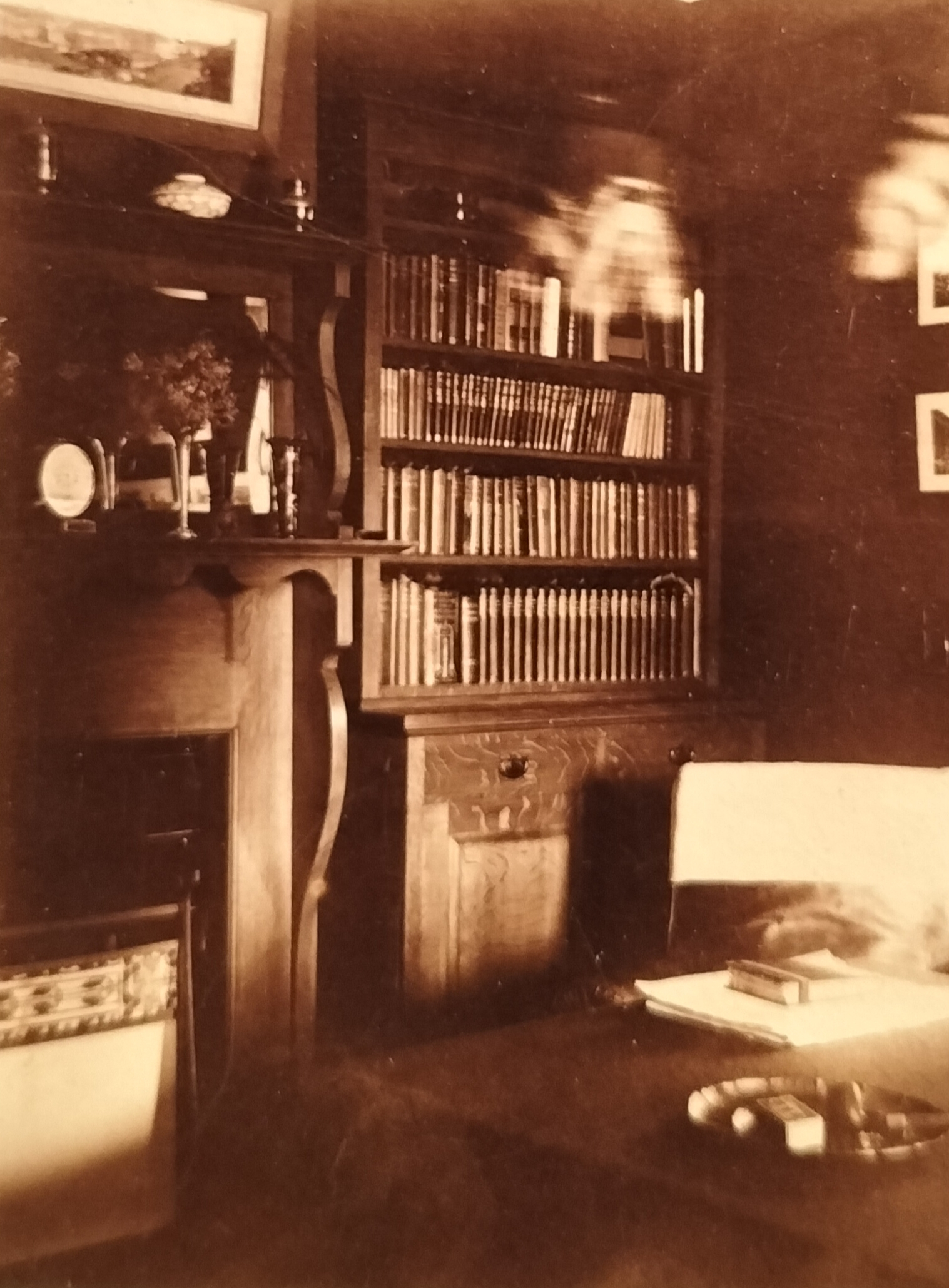 In the lounge with its elaborate wooden fireplace surround. Note the pipe on the desk as well as the unusual panoramic photograph on the left. This photograph in all probability is of East London. The ceiling lamp is assumingly blurred due to movement caused by wind.