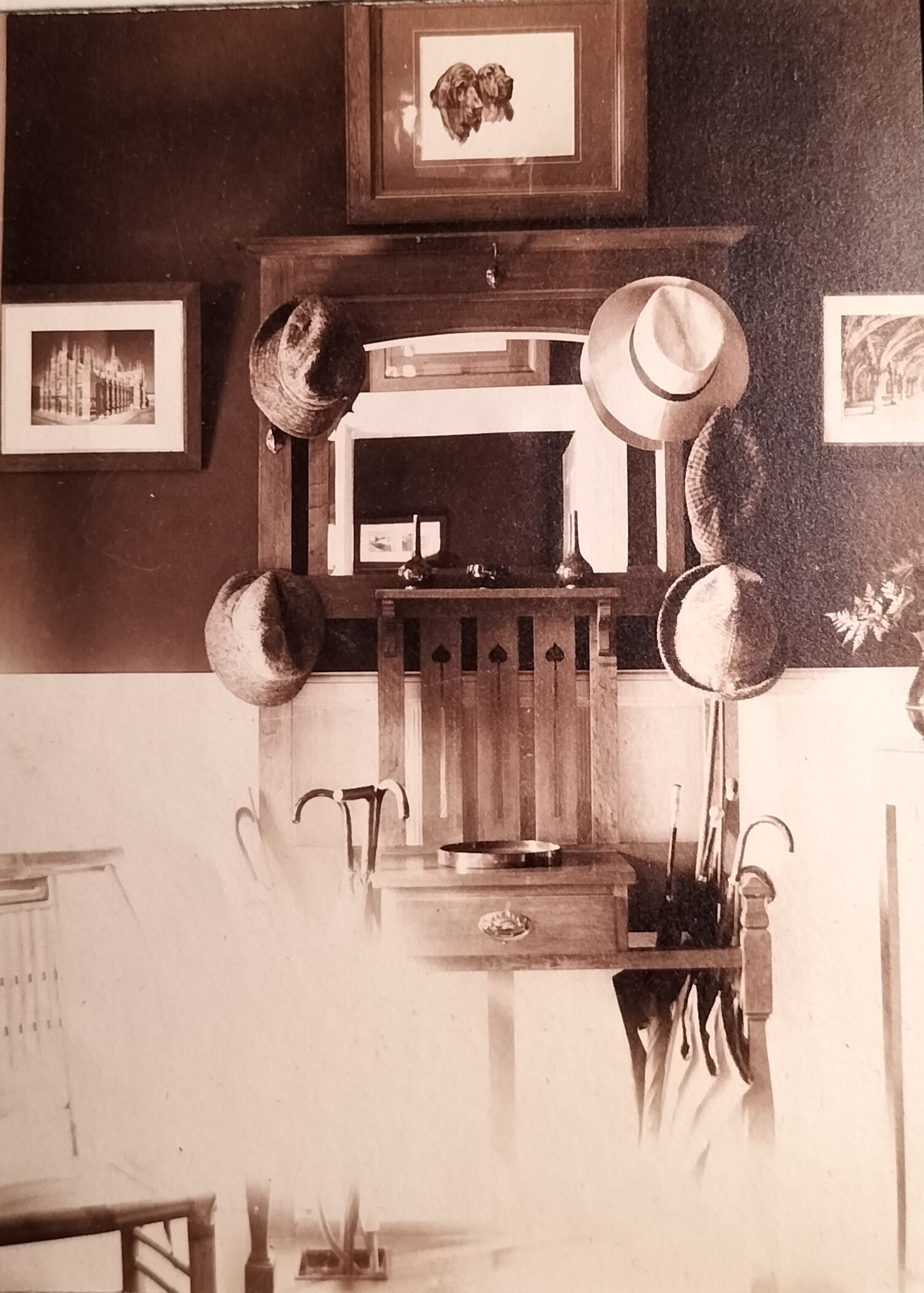A slightly overexposed photograph showing a chair and hall stand at the entrance hall with hats, walking sticks and umbrellas. Note the framed pictures of church structures as well as dogs.