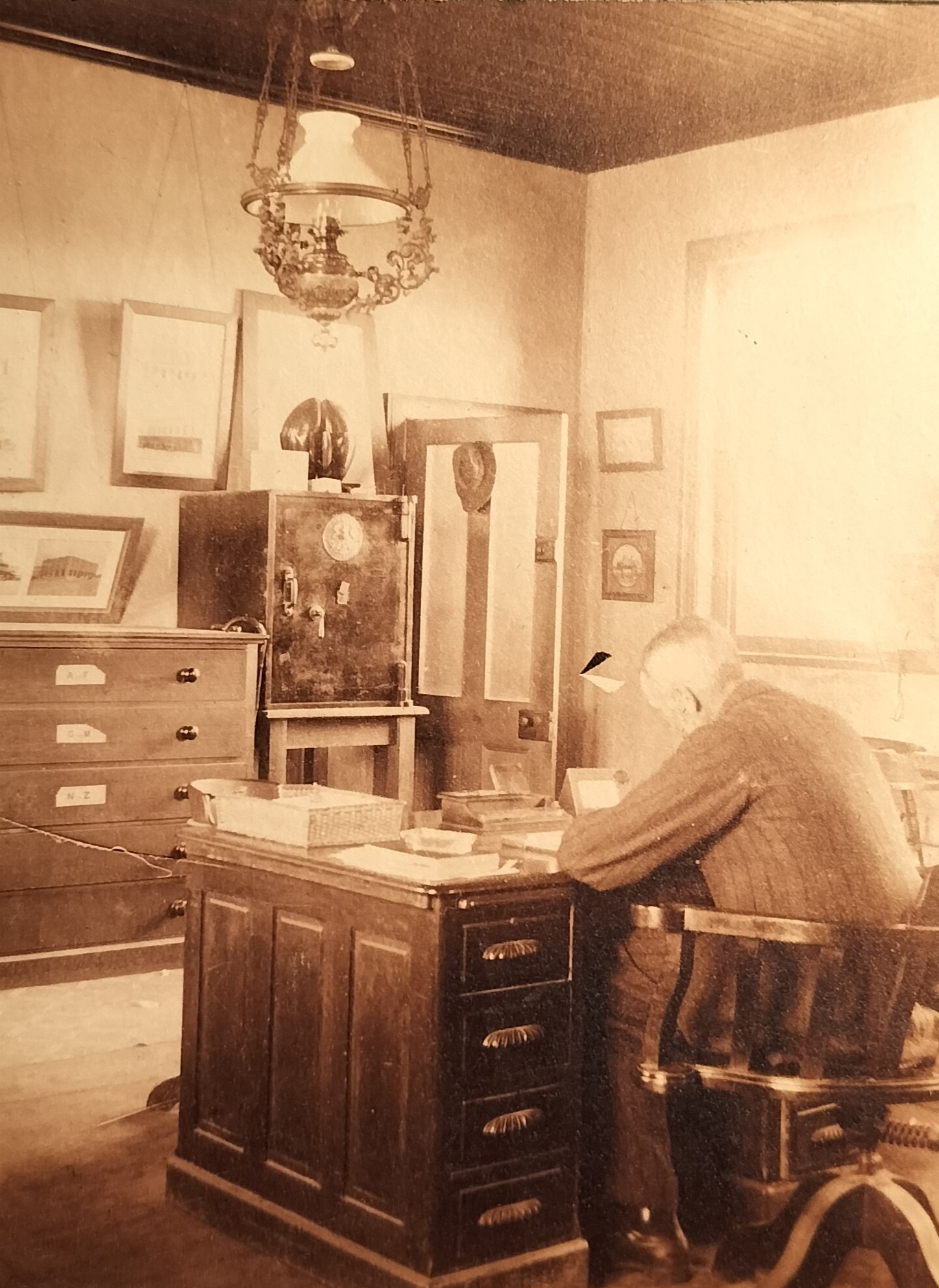 Another photograph of the male occupant in his study. The set of drawers suggests that he may have been an architect. This hypothesis is further supported by the two framed photographs of buildings hanging on the left. Note the Coco de Mer palm nut (Lodoicea maldivica) standing on top of the safe. There are also two walking canes visible between the set of drawers and the safe.